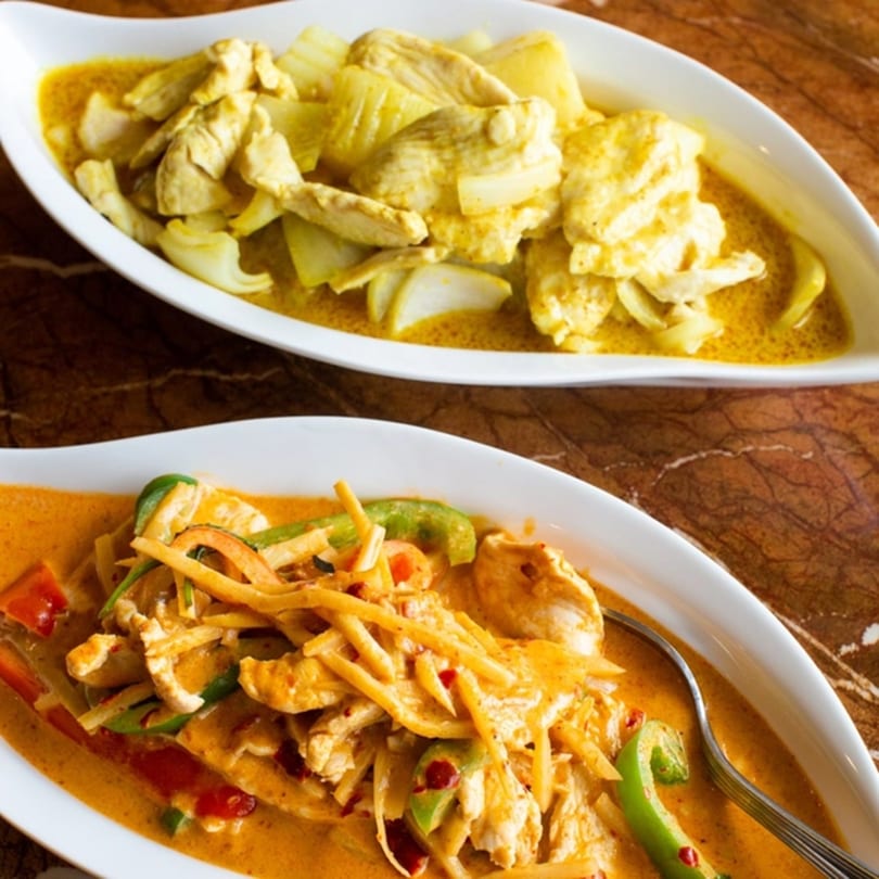 Two white oval dishes of Thai curries: yellow curry with chicken and onions, and red curry with chicken and vegetables.