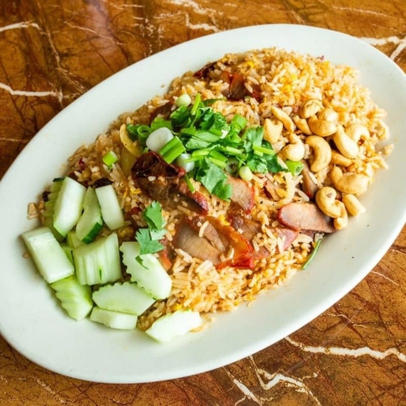 Thai fried rice with sliced pork, cashews, chopped green onions, and cucumber on a white plate.