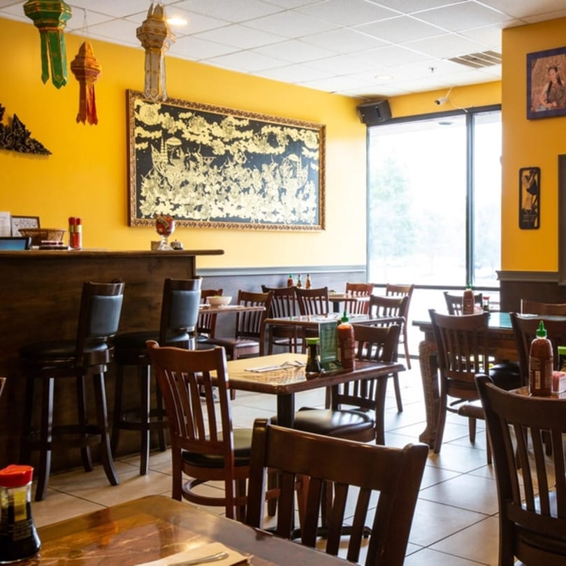 Interior of Thai Kitchen restaurant with yellow walls, dark wood tables, chairs, and a bar area.
