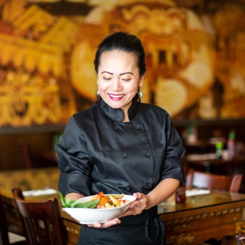 Smiling Thai Kitchen chef in black uniform holding a bowl of colorful Thai food.