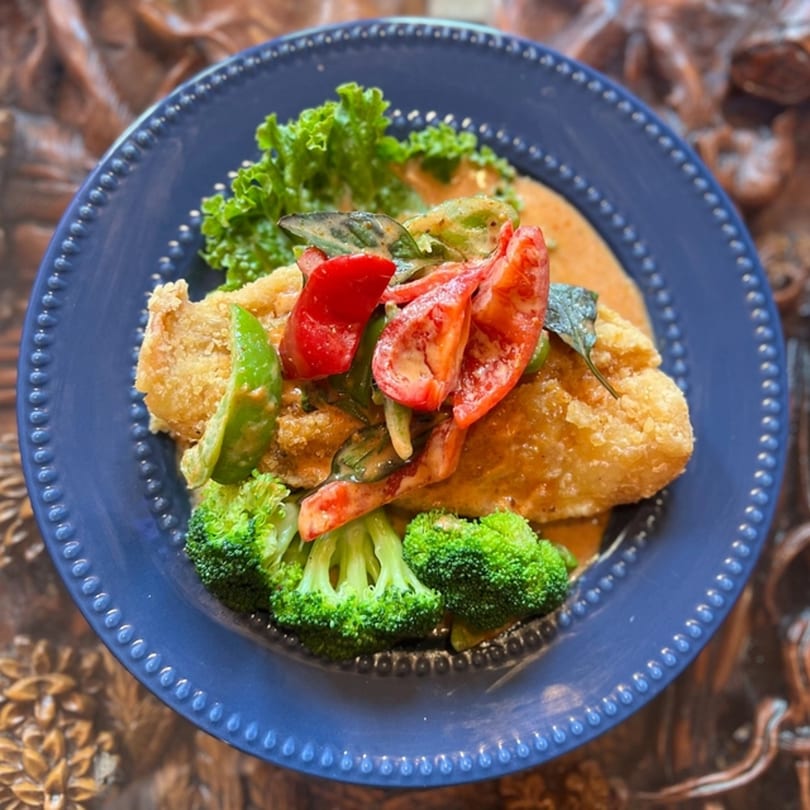 Crispy fried fish with red bell peppers, green vegetables, and a creamy sauce on a blue plate.
